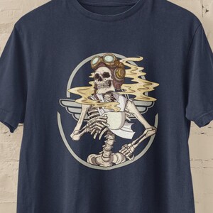 May include: A navy blue t-shirt with a graphic of a skeleton wearing a pilot's helmet and goggles, holding a cup of coffee. The skeleton is surrounded by a circle with wings and smoke coming from the coffee cup.