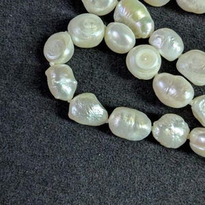 May include: A strand of white baroque pearls, with a slight iridescent sheen, arranged in a loose circle on a black background.
