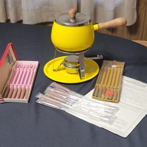 May include: A vintage fondue set with a bright yellow pot, a stainless steel stand, and a wooden handle. Includes a box of fondue forks and a box of fondue skewers. The set is on a dark blue surface.