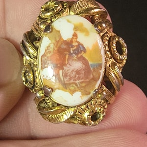May include: A gold-tone ring with a cameo featuring a woman in a blue dress. The cameo is surrounded by a decorative gold frame with leaves and flowers.
