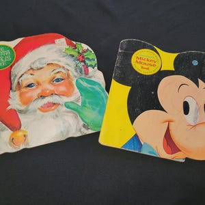 May include: Two vintage children's books. One is shaped like Santa Claus, with a red hat and green glove. The other is shaped like Mickey Mouse, with a yellow background and black ears. Both books are slightly worn.