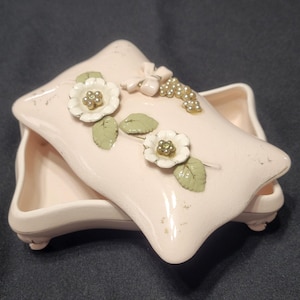 May include: A light pink ceramic jewelry box with a pillow-shaped lid. The lid is decorated with white flowers, green leaves, and a cluster of small, pearl-like beads. The box has a rectangular base and small feet.