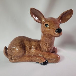 May include: A ceramic figurine of a fawn in a resting pose. The fawn has brown fur, large ears, and black hooves. The figurine is glazed with a glossy finish, giving it a polished appearance. The eyes are black and the nose is black.