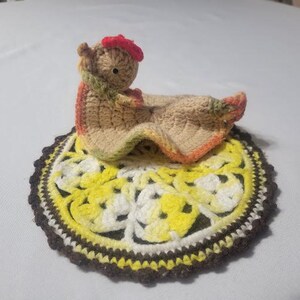 May include: A crocheted tan chicken-shaped lovey with a red comb and green accents, resting on a round, multi-colored crocheted doily featuring yellow, white, and dark brown yarn in a circular, floral-inspired pattern.