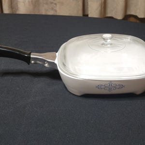 May include: White square casserole dish with a clear glass lid and black handle. The dish has a blue floral design on the front. The handle is black and attached to the dish with silver metal. The lid has a small knob.