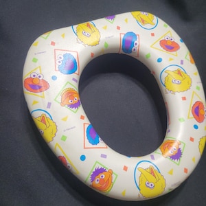 May include: A white Sesame Street potty training seat featuring colorful cartoon character graphics. The seat has a rounded shape with a central opening and is decorated with images of Elmo, Big Bird, and other characters.