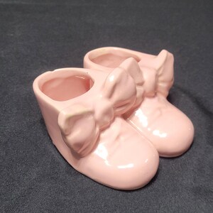 May include: A pair of small, pink ceramic baby shoes with large bow accents. The shoes are a light pastel pink color and have a glossy finish. The shoes are designed to be decorative.