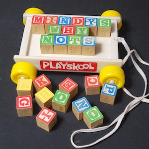 May include: A vintage Playskool pull toy with a white cart, yellow wheels, and a white pull string. The cart holds wooden blocks with letters and the words "MINDYS", "WHY", and "NOTS". Additional blocks are scattered around.