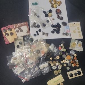 May include: A collection of various buttons in different sizes, colors, and materials. Some buttons are on cards, others in small plastic bags. Colors include black, white, gold, brown, and green. Some cards read "Buttons" and "le chic".