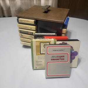 May include: A wooden box with a metal handle, filled with cassette tapes. A "Realistic" brand cleaner and demagnetizer box is in front. The box is brown with a dark brown lid. The tapes are in various colors.