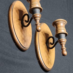 May include: Two wooden wall sconces with a dark brown finish. Each sconce features a light brown, oval-shaped backplate, a decorative black metal arm, and a wooden candle holder. The sconces are designed to hold candles and add a warm ambiance to any room.