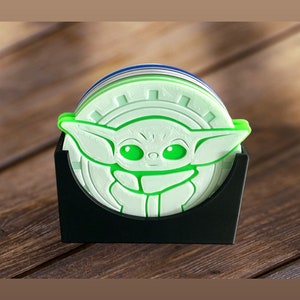 Puede incluir: Un juego de seis posavasos verdes y blancos con un contorno verde de un personaje de Star Wars: The Mandalorian. Los posavasos están en un soporte negro.
