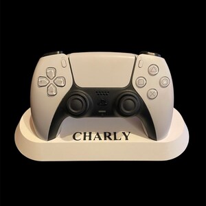 Peut inclure: Une manette de jeu PlayStation 5 blanche avec des boutons et des gâchettes noirs, reposant sur un support blanc avec le nom "CHARLY" imprimé dessus.