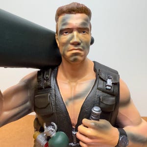 Puede incluir: Una detallada figura de un soldado musculoso con pintura de camuflaje en la cara, que lleva un chaleco táctico negro y lleva un objeto grande y verde oscuro. La figura tiene una expresión seria y un reloj de pulsera.