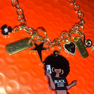 May include: A gold chain bracelet with multiple charms, including a black and white star, a black heart with gold trim, a black flower with a white pearl center, a gold tag with the word "BRAVE" on it, a gold tag with the word "BEAUTIFUL" on it, and a charm of a black woman with an afro holding a phone with the text "BLACK. NO SUGAR. NO CREAM."