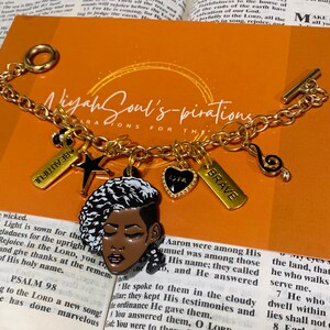 May include: A gold chain bracelet with charms, including a black and white portrait of a woman, a star, a music note, a heart with the word "Love" on it, and a tag that says "Brave". The bracelet also has a tag that says "Beautiful".