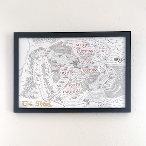 Puede incluir: Un mapa en blanco y negro de La Comarca, una región ficticia de los libros de El Señor de los Anillos, con texto rojo que destaca las diferentes áreas de La Comarca: Norte de la Comarca, Sur de la Comarca, Este de la Comarca y Oeste de la Comarca.