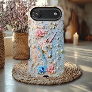 May include: A phone case featuring a detailed, embossed design of a unicorn with a pink horn, surrounded by flowers and pearls. The case has a light blue background and is set on a woven mat.