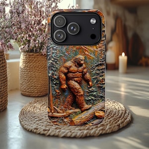 May include: A phone case featuring a detailed relief of a Bigfoot figure in a forest setting. The case has a textured, brown and green design with a black camera module. The Bigfoot is depicted walking on a log, with trees and mountains in the background.