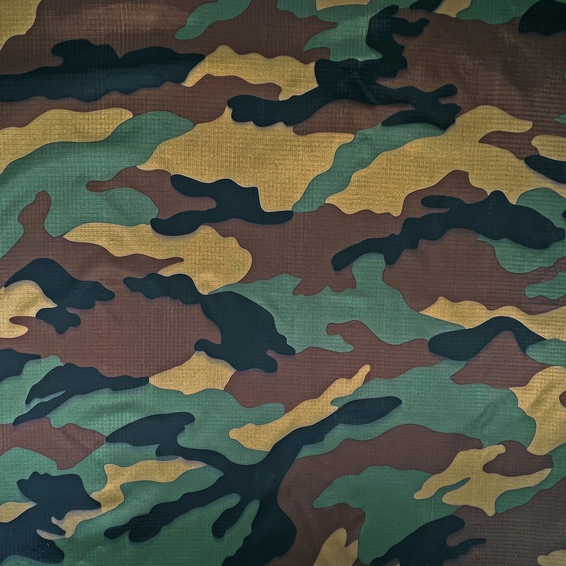May include: Camouflage pattern fabric featuring a mix of green, brown, black, and tan irregular shapes. The fabric has a textured appearance, with a subtle grid-like pattern visible throughout. This military-inspired design is suitable for various applications.