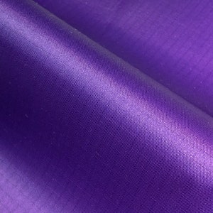 May include: Close-up of a folded piece of vibrant purple fabric. The material has a subtle sheen, with visible woven texture and slight variations in color. The fabric is smooth and appears to be lightweight.