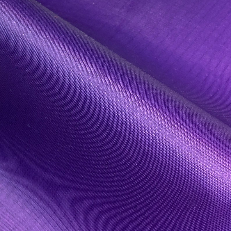 May include: Close-up of a folded piece of vibrant purple fabric. The material has a subtle sheen, with visible woven texture and slight variations in color. The fabric is smooth and appears to be lightweight.
