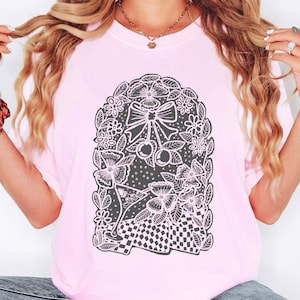 Może przedstawiać: Jasnoróżowa koszulka z czarno-białym wzorem graficznym. Projekt zawiera wieniec kwiatowy z kokardą, wiśniami i kieliszkiem do koktajli. Dół wzoru ma wzór w szachownicę.