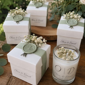 Può includere: Scatole regalo quadrate bianche con nastri verde salvia, sigilli in ceralacca con motivo a foglie e fiori secchi di gipsofila. Ogni scatola ha un'etichetta con scritto Olivia & Daniel 14.09.2026. Una candela in vetro riporta le lettere O e D.