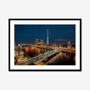 May include: A framed photograph of a cityscape at dusk. The image features a river with bridges, illuminated buildings, and a tall tower. The sky transitions from dark blue to black, with city lights reflecting on the water.