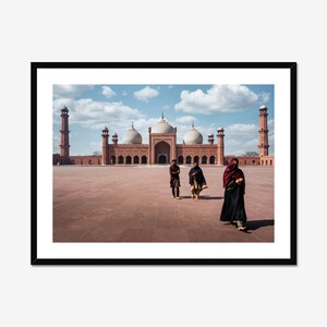 May include: A red sandstone mosque with three domes and two minarets. People are walking in the courtyard in front of the mosque.