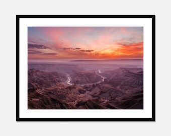 Può includere: Una vista panoramica di un canyon con un fiume che serpeggia attraverso di esso. Il cielo è di un arancione e rosa vibrante, creando un drammatico tramonto sul paesaggio.