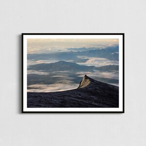 May include: A framed landscape photograph featuring a mountain peak in the foreground, with a vast expanse of mountains and clouds in the background. The sky transitions from a soft yellow to a pale blue. The image is framed in a black border.