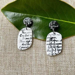 Op de afbeelding: Een paar handgemaakte oorbellen. Elke oorbel heeft een witte, langwerpige hanger met zwarte tekst en een zwarte, cirkelvormige bovenkant met witte accenten. De tekst lijkt een gedicht of citaat te zijn.