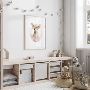 K&ouml;nnte beinhalten: Ein gerahmter Wandkunstdruck mit einer Aquarellillustration eines jungen Rehs mit gro&szlig;en Ohren, geschm&uuml;ckt mit einem Kranz aus zarten rosa und pfirsichfarbenen Blumen und gr&uuml;nen Bl&auml;ttern, zentriert in einem feinen Blumenkranz.