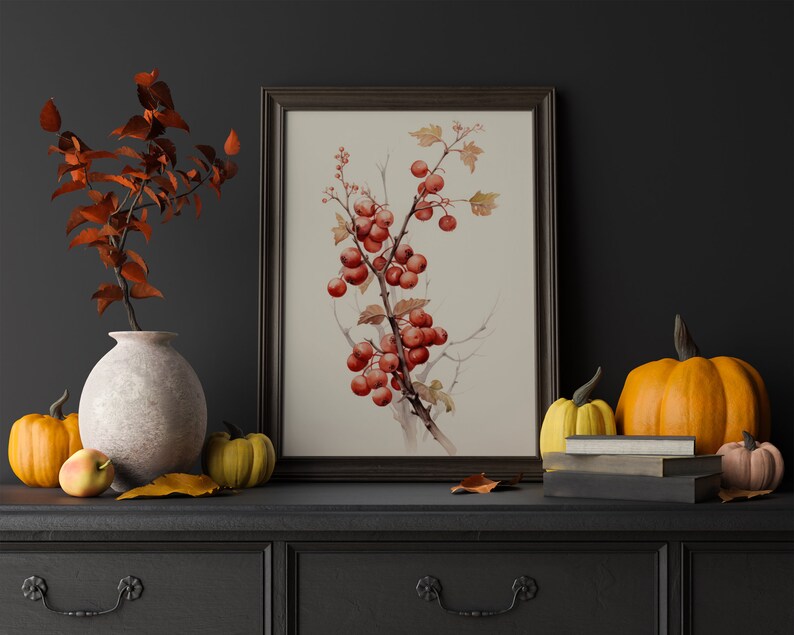 May include: A framed botanical print of a branch with red berries against a white background. The print is on a dark grey dresser with a vase of fall leaves, pumpkins, and books.