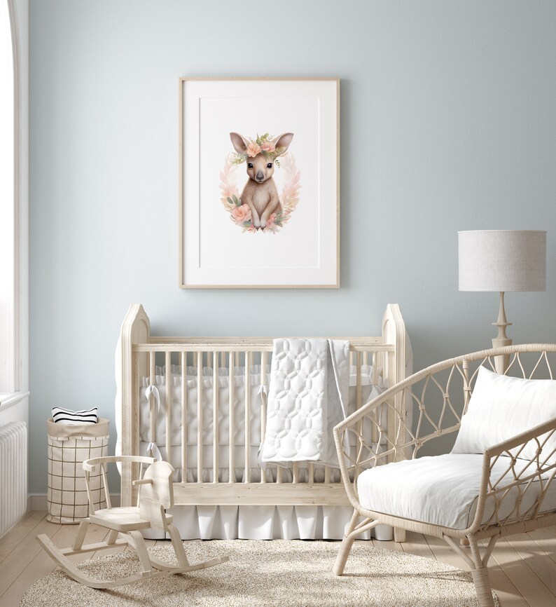 K&ouml;nnte beinhalten: Ein gerahmter Kunstdruck mit einer Aquarellillustration eines jungen K&auml;ngurus, das in einem Kranz aus zartfarbenen Blumen sitzt. Das K&auml;nguru hat gro&szlig;e Ohren und ist von rosa und pfirsichfarbenen Bl&uuml;ten sowie gr&uuml;nem Blattwerk umgeben.
