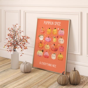 May include: A framed orange print with the words "PUMPKIN SPICE & EVERYTHING NICE" and illustrations of smiling pumpkins. A white vase with orange branches and two decorative pumpkins are in front of a white wall.