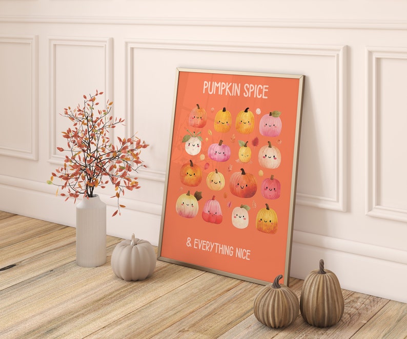 May include: A framed orange print with the words "PUMPKIN SPICE & EVERYTHING NICE" and illustrations of smiling pumpkins. A white vase with orange branches and two decorative pumpkins are in front of a white wall.