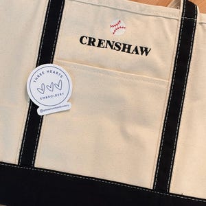 May include: Cream-colored tote bag with black accents, featuring the embroidered word "CRENSHAW" and a baseball graphic. The bag has a front pocket and black handles. A sticker with the text "THREE HEARTS EMBROIDERY" is attached.