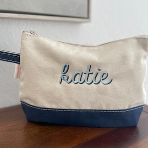 May include: A beige canvas cosmetic bag with a navy blue base and handle. The name "Katie" is embroidered in light blue script across the front. The bag has a zip closure and a small tag on the side.