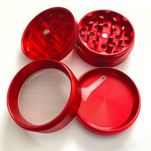 Puede incluir: Un molinillo de hierbas de metal rojo de cuatro piezas desmontado en sus componentes individuales. El conjunto incluye dos secciones de tapa con dientes afilados, una secci&oacute;n central con una malla met&aacute;lica fina y una bandeja base con una peque&ntilde;a esp&aacute;tula de pl&aacute;stico blanco.