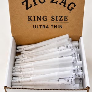Peut inclure: Une boîte en carton contenant de nombreux cônes en papier blanc pré-roulés. Le couvercle ouvert porte le texte noir ZIG ZAG, KING SIZE et ULTRA THIN. Chaque cône est marqué du logo Zig Zag et des mots REMOVE BEFORE USE.