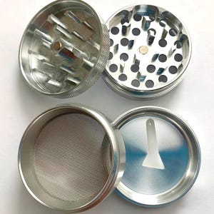 Puede incluir: Un molinillo de hierbas de metal plateado de cuatro piezas mostrado desmontado. Las dos secciones superiores tienen dientes afilados en forma de diamante, la tercera contiene una malla met&aacute;lica fina y la base incluye una peque&ntilde;a esp&aacute;tula blanca.