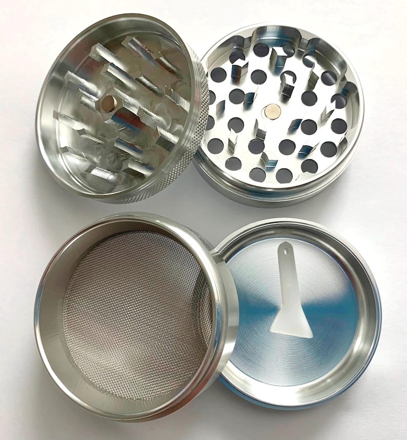 Puede incluir: Un molinillo de hierbas de metal plateado de cuatro piezas mostrado desmontado. Las dos secciones superiores tienen dientes afilados en forma de diamante, la tercera contiene una malla met&aacute;lica fina y la base incluye una peque&ntilde;a esp&aacute;tula blanca.