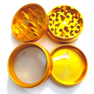 Puede incluir: Un molinillo de hierbas de metal dorado de cuatro piezas desmontado en sus componentes individuales. El conjunto incluye dos secciones de trituraci&oacute;n con dientes afilados, una secci&oacute;n con malla fina, una base s&oacute;lida y una peque&ntilde;a esp&aacute;tula de pl&aacute;stico blanco.