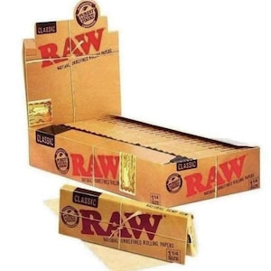 Puede incluir: Una caja de papeles de liar RAW Classic de tamaño 1 1/4 naturales sin refinar. La caja es marrón con un logotipo RAW rojo y el texto "Authentic Purest Natural Fibers". La caja está abierta y contiene varios paquetes de papeles de liar. También hay un solo paquete de papeles de liar RAW Classic de tamaño 1 1/4 naturales sin refinar sobre una superficie marrón.