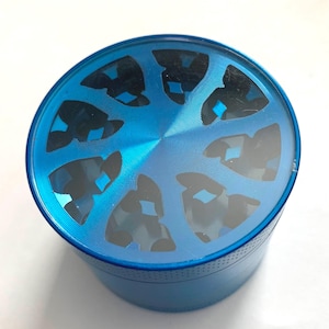 Puede incluir: Un molinillo de hierbas cil&iacute;ndrico de metal azul con una tapa superior transparente. La tapa presenta un patr&oacute;n circular de ocho recortes negros de forma triangular dispuestos alrededor de un punto central.
