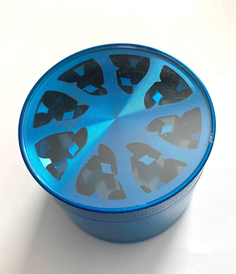 Puede incluir: Un molinillo de hierbas cil&iacute;ndrico de metal azul con una tapa superior transparente. La tapa presenta un patr&oacute;n circular de ocho recortes negros de forma triangular dispuestos alrededor de un punto central.