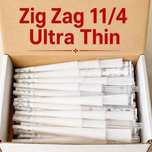May include: A cardboard box filled with white Zig Zag 1 1/4 Ultra Thin pre-rolled cones. The interior lid of the box features the red text Zig Zag 1 1/4 Ultra Thin. Each cone includes a paper filter tip with the Zig Zag logo printed on it.