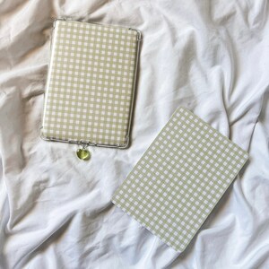 May include: Two beige and white gingham patterned cases, one with a silver frame and a green heart charm. The cases are on a white fabric surface.
